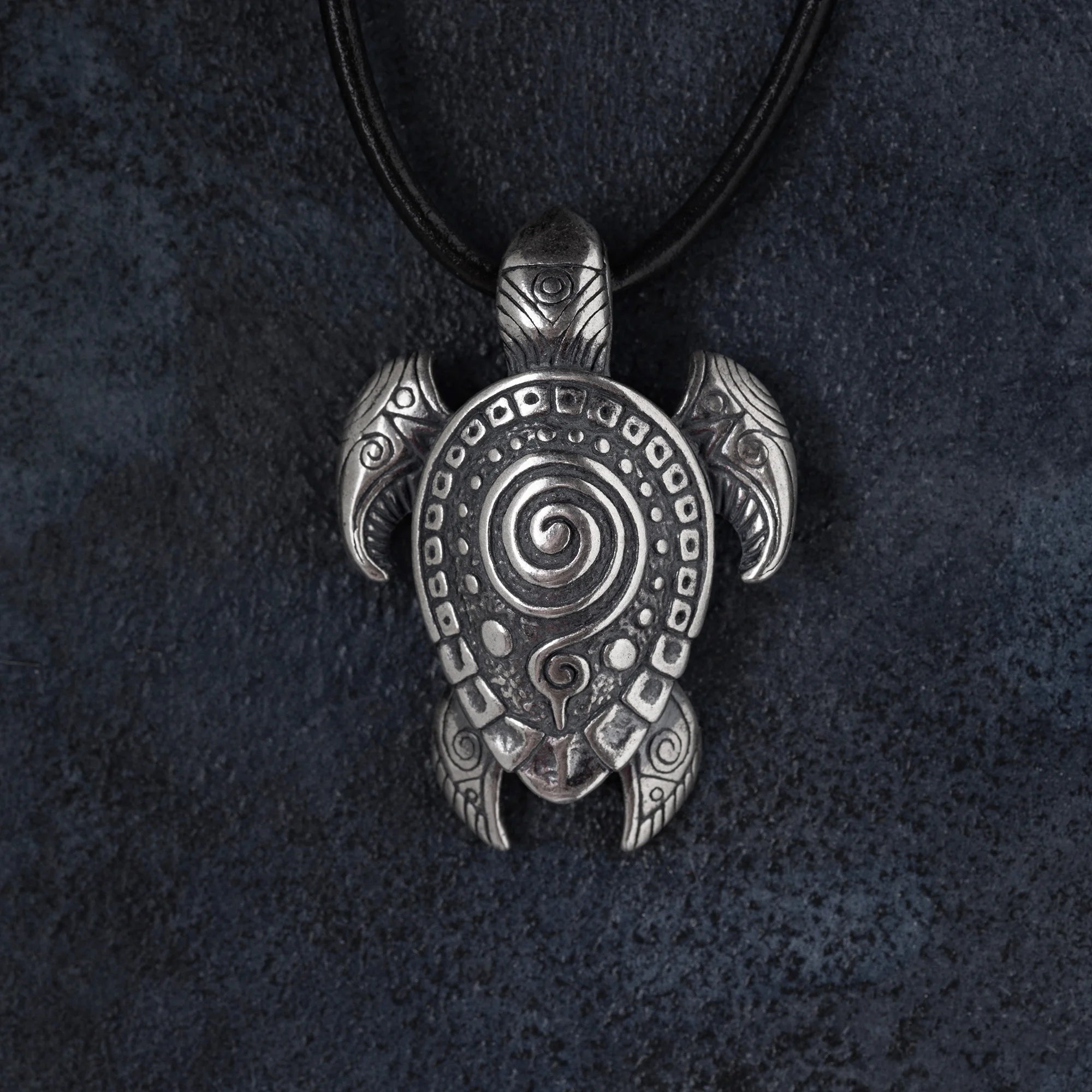 An engraved silver turtle pendant on a black cord necklace, displayed against a dark textured background.