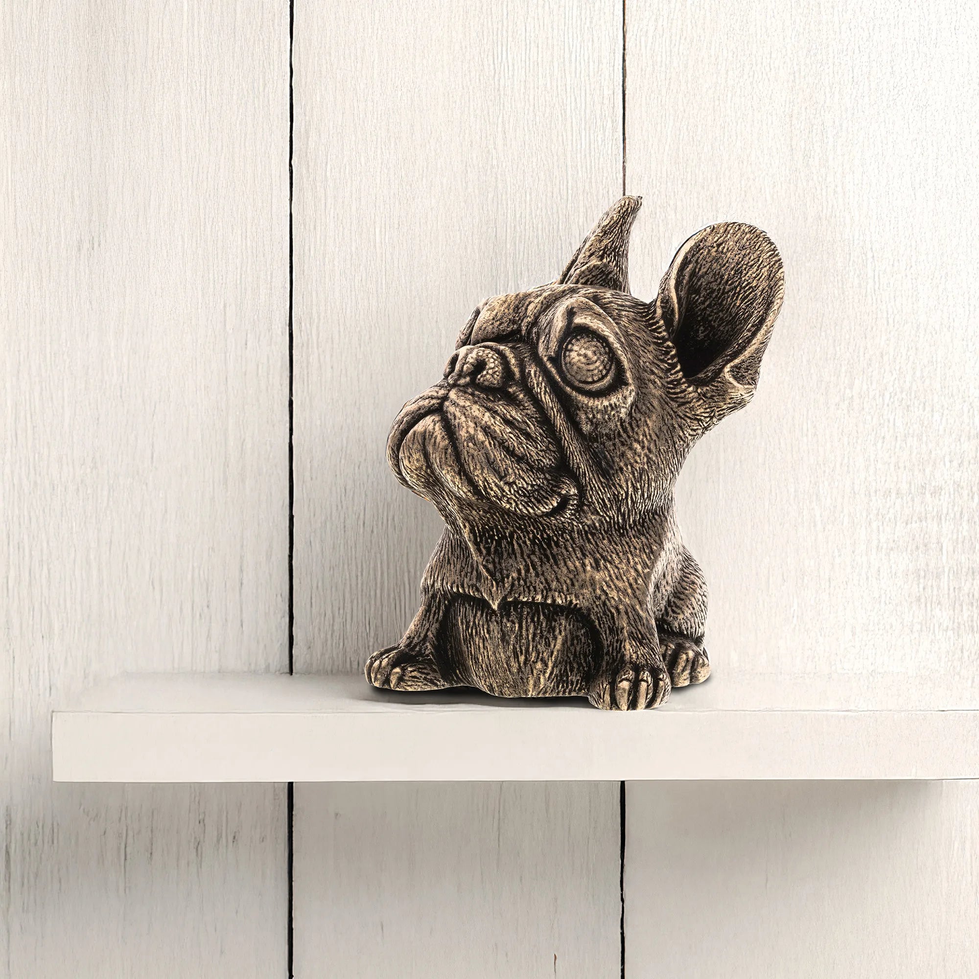 A small French bulldog figurine with a textured, metallic bronze finish, sitting on a white shelf against a white wood-paneled background.