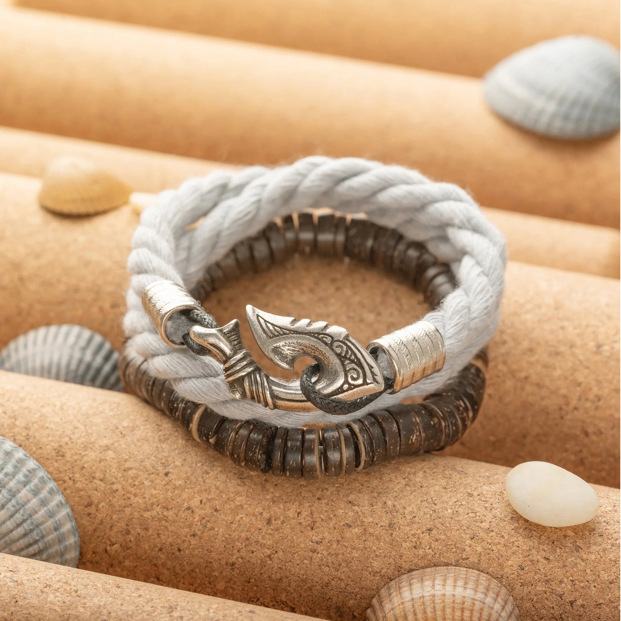 A braided rope bracelet with a Viking-style axe clasp, layered over dark metallic beaded strands, displayed on a cork jewelry organizer with seashells.