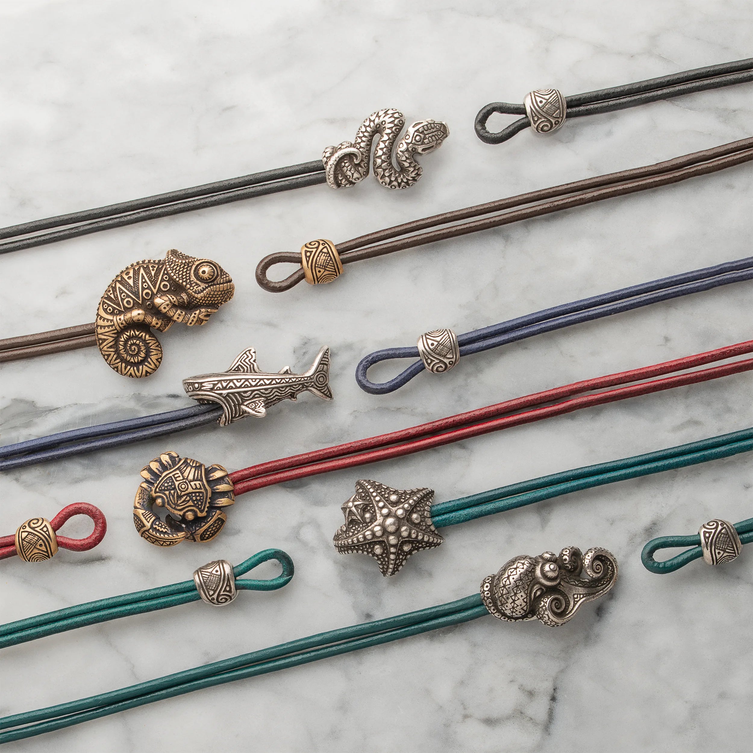 A flat-lay display of multiple multi-strand leather cord jewelry pieces with various animal and nature charms, including a chameleon, snake, shark, starfish, lion, and cloud, arranged on a white marble background.
