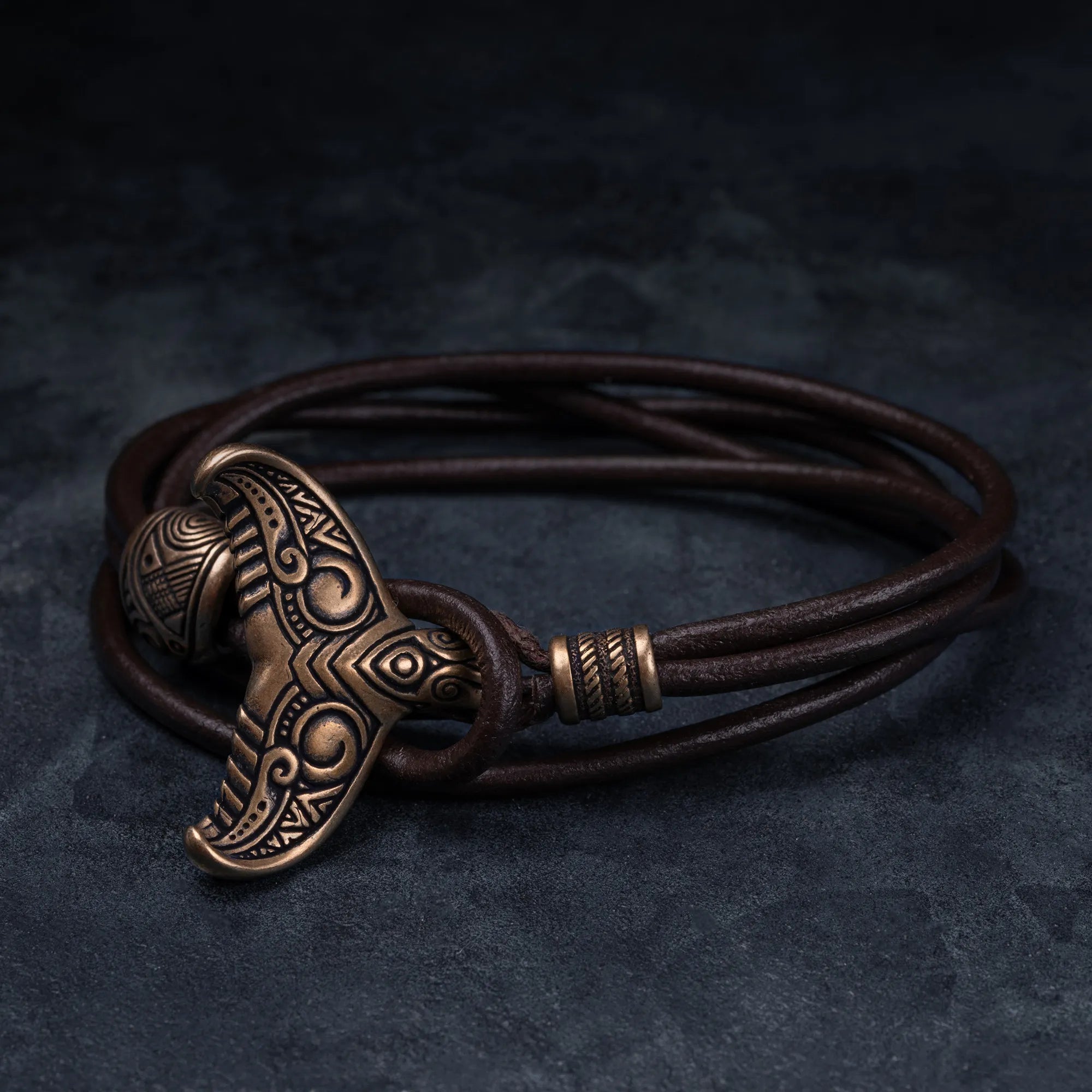 A dark brown leather wrap bracelet featuring a detailed bronze Viking-style hammer pendant, displayed on a dark textured background.