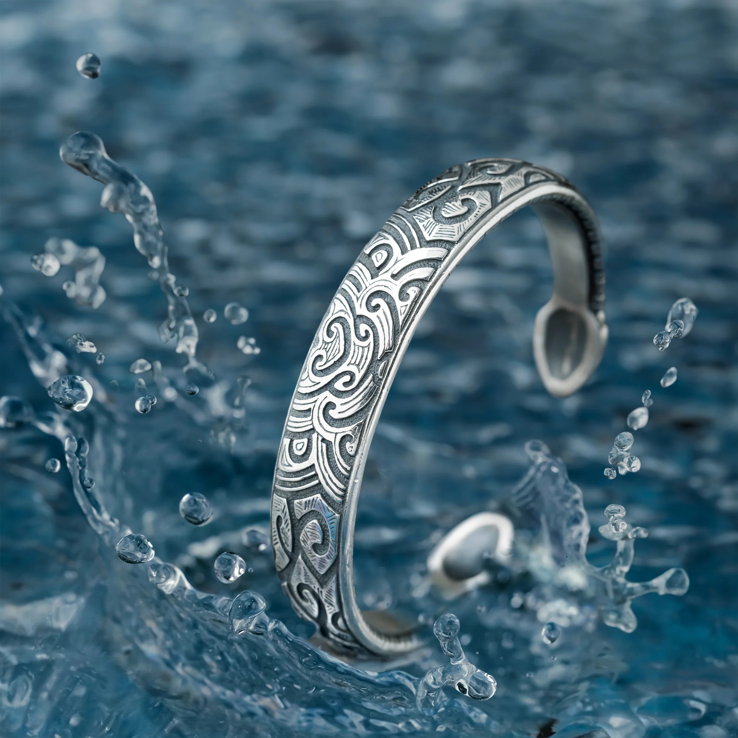 A silver-toned open cuff bracelet with an engraved wave or tribal pattern, shown partially submerged in splashing blue water.