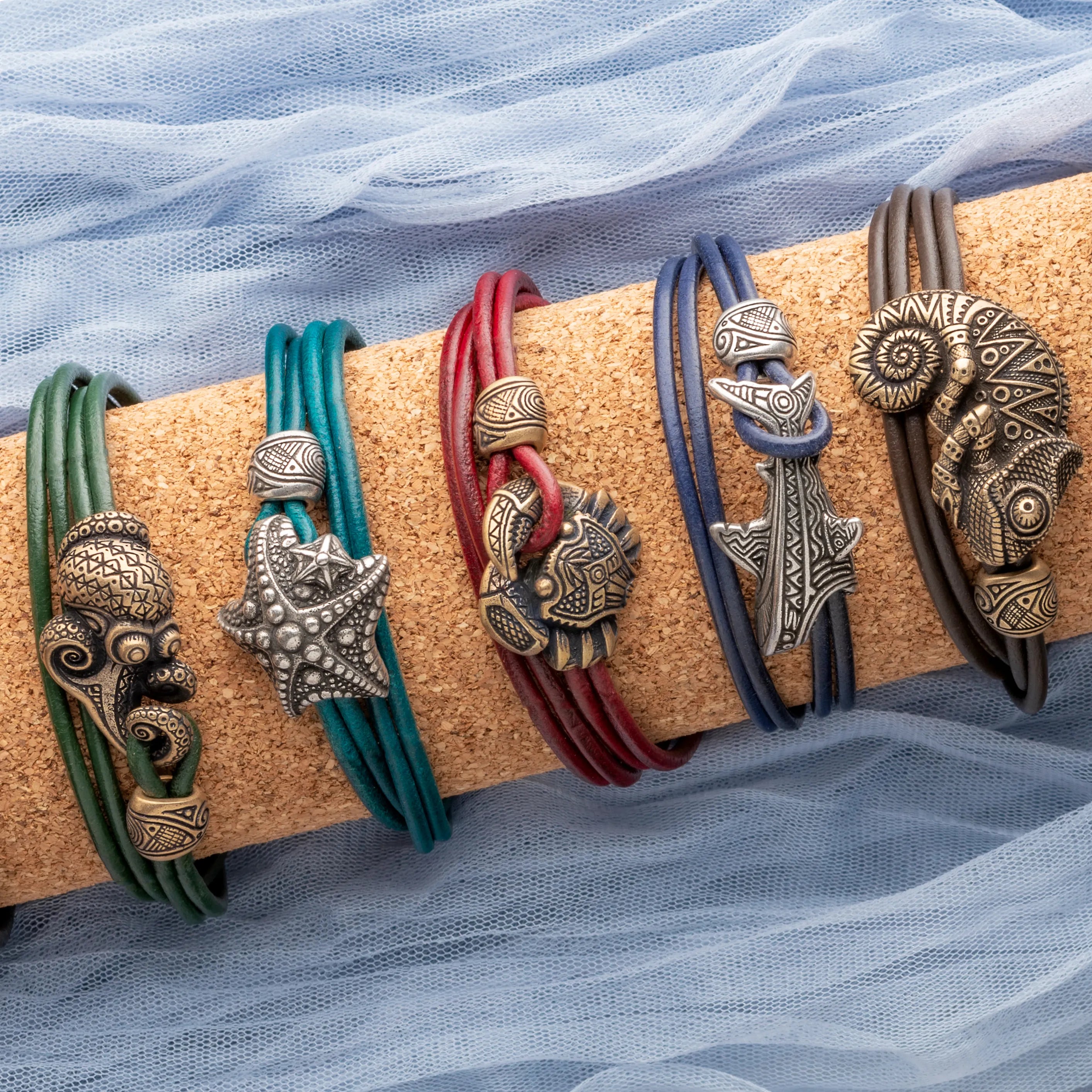 Five leather wrap bracelets with sea-themed metal charms are displayed on a cork roll wrapped in sheer blue fabric. The charms include an octopus, a starfish, a lion, a shark, and a ram.