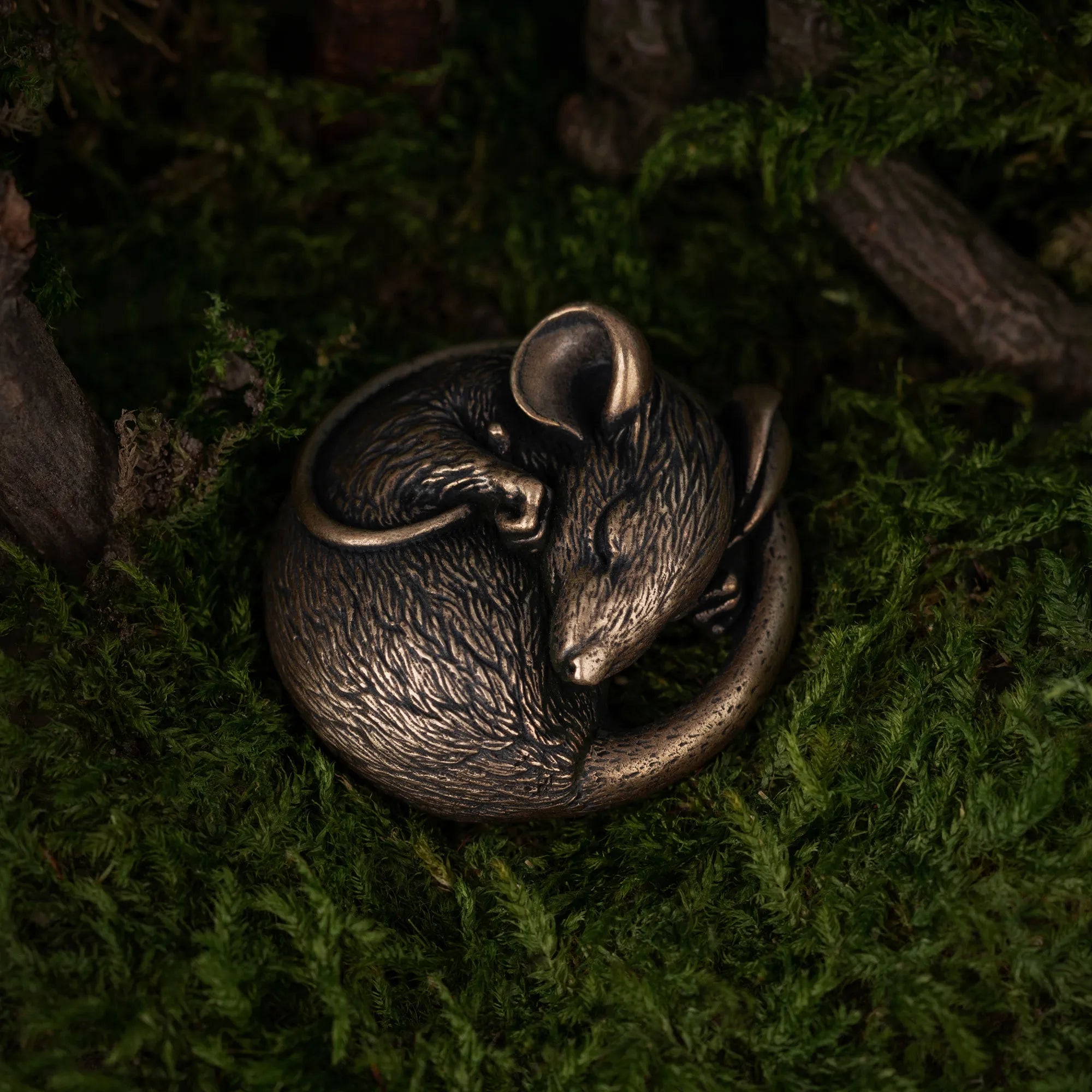 A small metallic mouse figurine curled up in a sleeping pose, resting on dark green moss with blurred natural wood elements in the background.