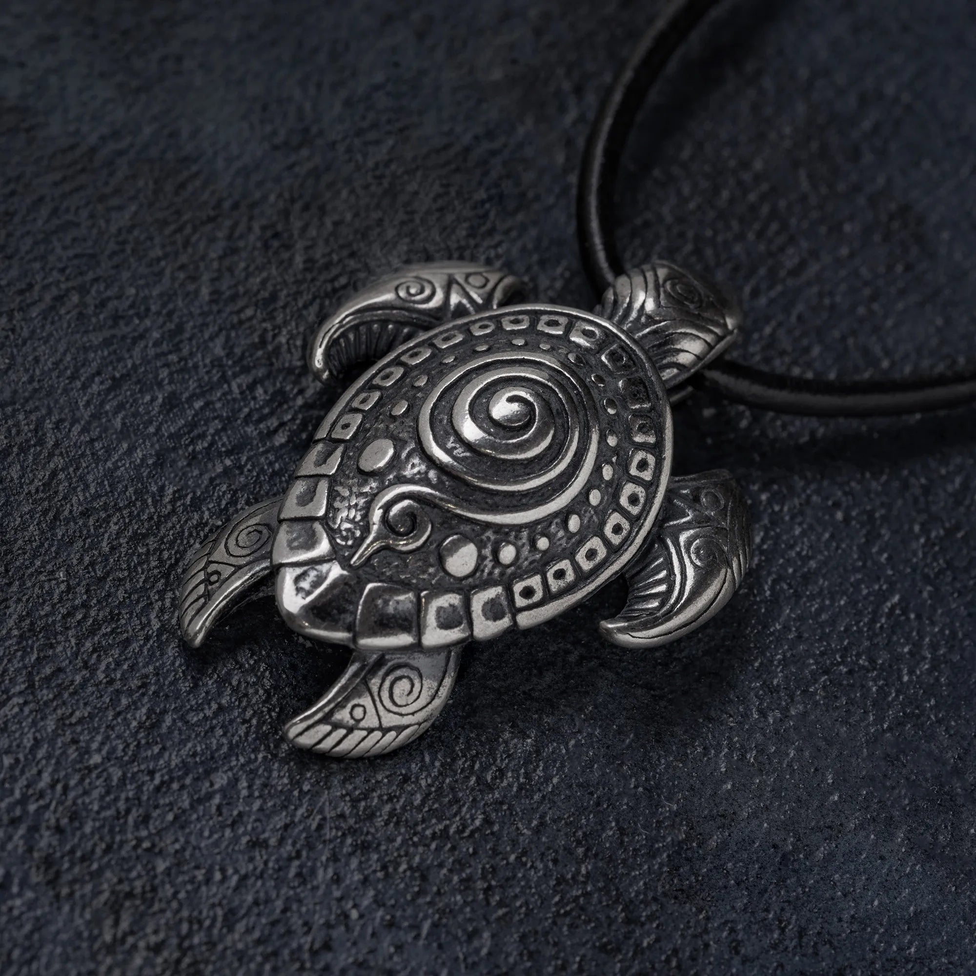 A silver-toned metal turtle pendant with intricate engraved patterns, hanging from a black cord against a dark textured background.