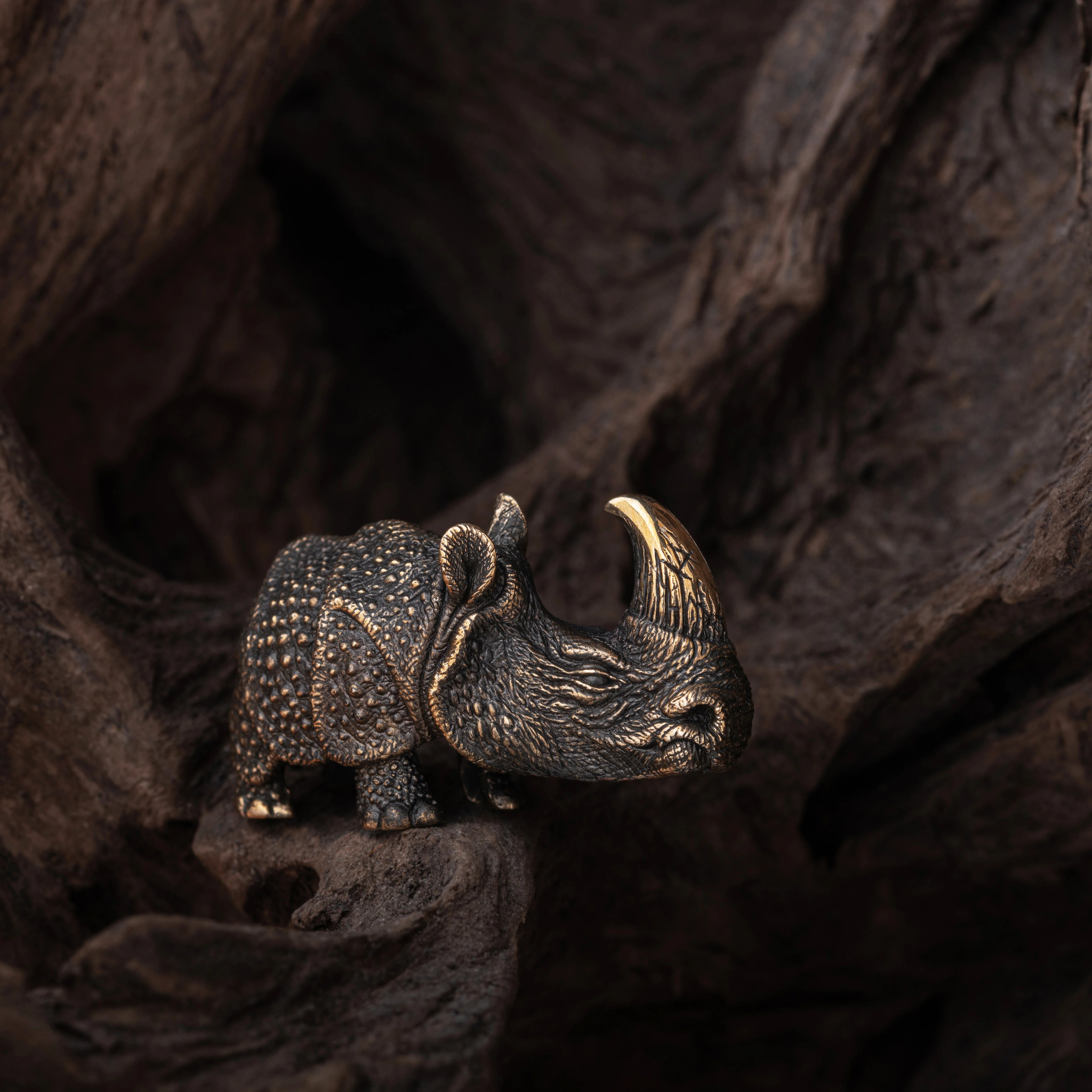 A small rhinoceros figurine with textured dark metallic scales and a prominent curved horn, posed on a dark wooden surface against a rustic wood background.